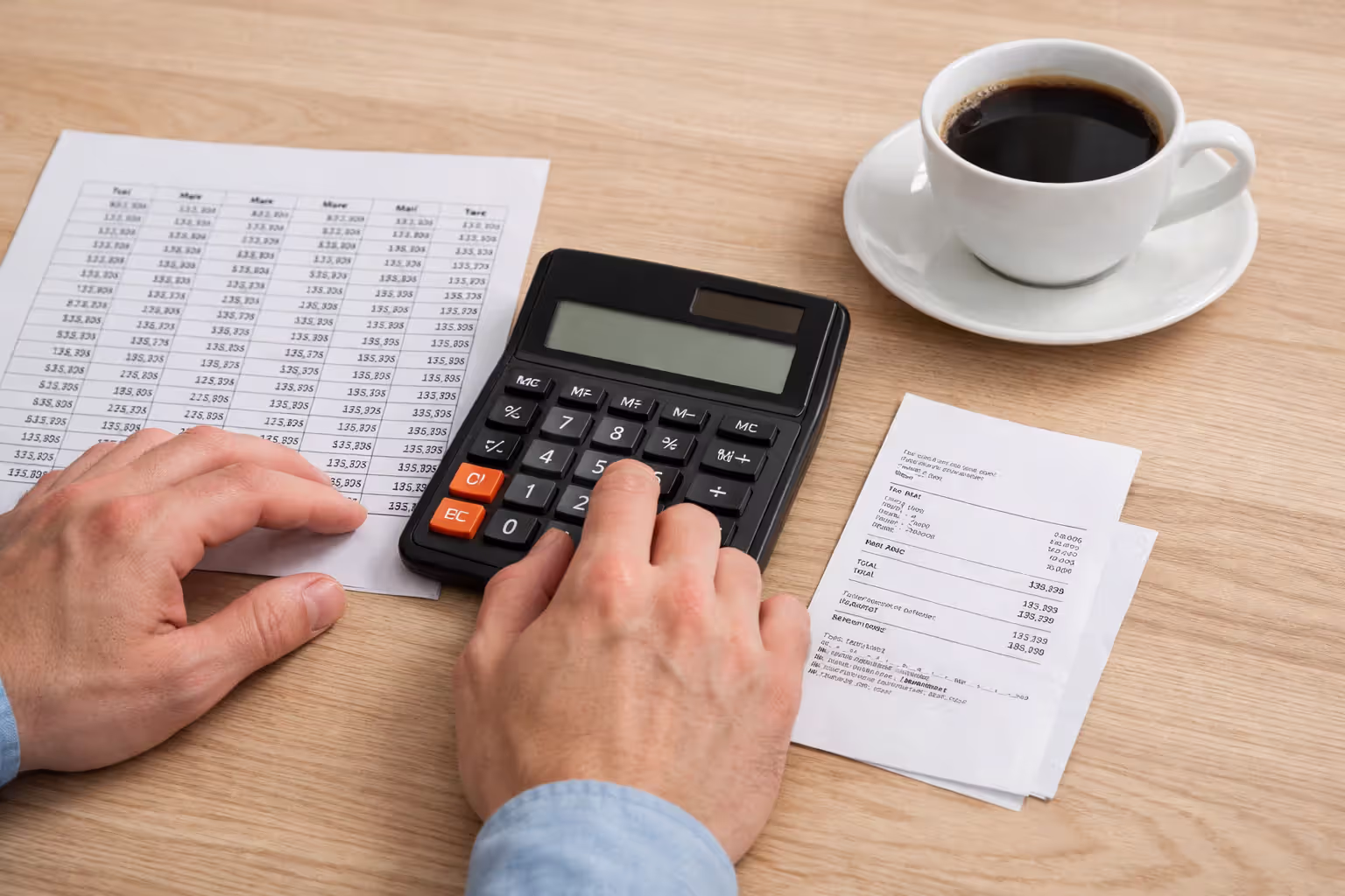 Close-up of hands using a calculator next to a printed payment schedule and coffee cup on a wooden desk for student loan payment calculation