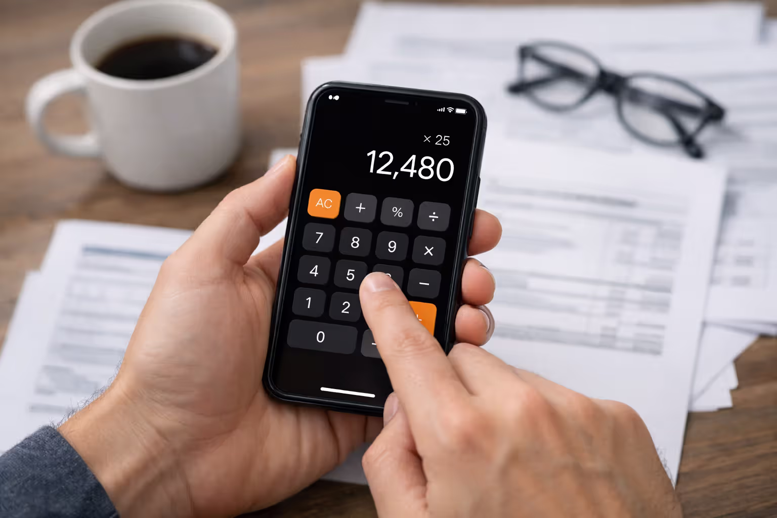 Hands holding smartphone with calculator app over blurred desk with tax documents and glasses